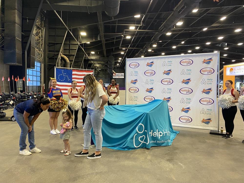 Emberly talks to Kelly L Green as Kelly prepares to unveil Emberly’s new adaptive bike, covered by a large teal Help Hope Live banner, at the Abilities Expo Phoenix.