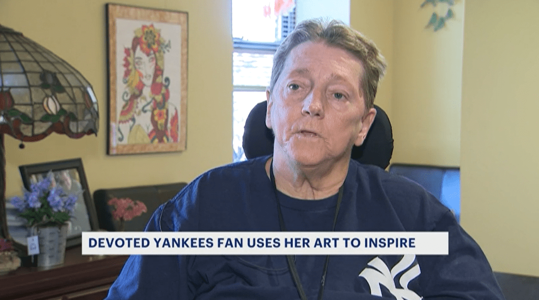 Help Hope Live client Jane Koza explains on the news how she uses her artwork to inspire others and raise funds. Jane has short sandy hair and light skin. Her colorful artwork is visible on the wall behind her. A ticker caption reads Devoted Yankees Fan Uses Her Art to Inspire.