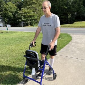 Double lung transplant recipient Terry Freeman is outdoors on a sidewalk walking with a blue walker. He has light skin, short gray hair, sunglasses, and a gray t-shirt that reads Clayton Fear Farm.
