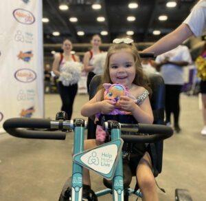 3-year-old Emberly smiles holding a doll as she sits on her new adaptive bike. She is at the Abilities Expo Phoenix.